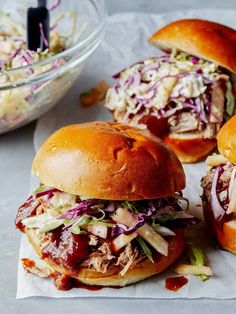 three pulled pork sandwiches with coleslaw slaw on the side and a bowl of coleslaw in the background