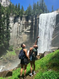 two women standing in front of a waterfall pointing up at the sky with their hands