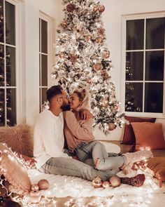 a man and woman sitting on the floor in front of a christmas tree with lights