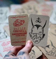 a person holding up two cards with ink drawings on them, one has a demon's head and the other has a devil's head