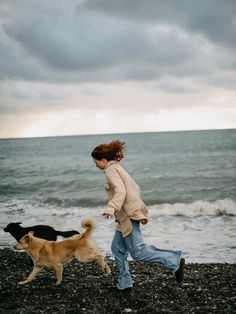 a woman walking on the beach with two dogs running behind her in front of the ocean