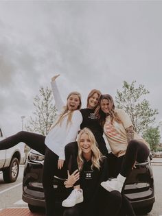 three girls are posing on the hood of a car in a parking lot with their arms up