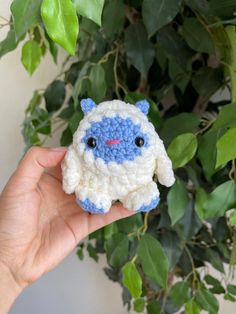 a hand holding a small crocheted blue and white sheep ornament in front of a potted plant