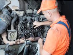 a man in an orange shirt working on the engine of a vehicle with other workers around him