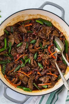 a pot filled with meat and vegetables on top of a white table next to silver spoons