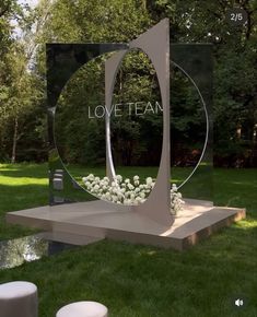 a sculpture in the middle of a grassy area with white flowers and trees behind it