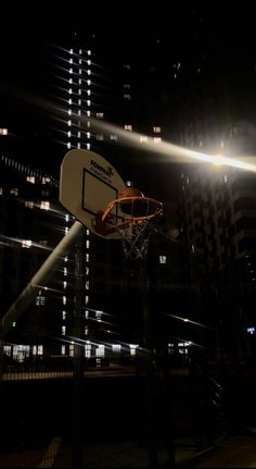 a basketball hoop in the air at night with lights shining down on buildings behind it