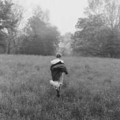 black and white photograph of a person running in a field with trees behind them on an overcast day