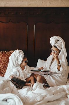 two women in white robes sitting on a bed reading a book and talking on the phone