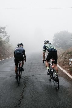 two bicyclists are riding down the road on a foggy day