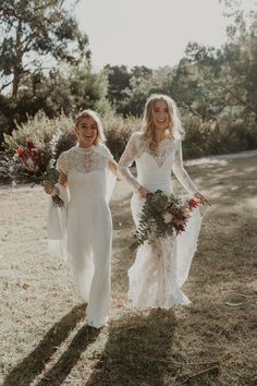 two women in white dresses are walking through the grass with flowers on their heads and holding bouquets