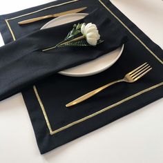 a place setting with black napkins, gold forks and a white flower on it