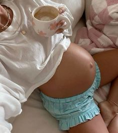 a pregnant woman laying in bed with her stomach exposed and holding a cup of coffee