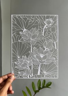 a hand holding up a piece of paper with white flowers on it and a plant in the background