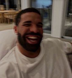 a man with a beard sitting on a couch smiling at the camera while wearing a white t - shirt