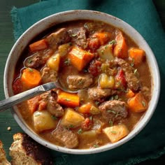 a bowl of stew with meat, potatoes and carrots on a green cloth next to bread