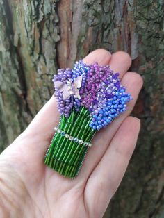 a hand holding a purple and blue flower brooch in front of a tree trunk