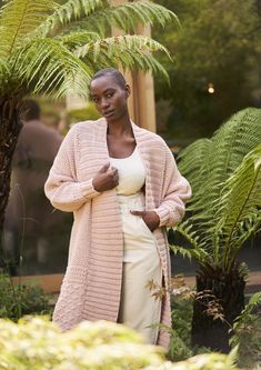 a woman in a white dress and pink cardigan standing next to some plants with her hand on her hip