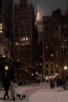 people are walking in the snow on a city street with tall buildings and lights at night