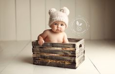 a baby is sitting in a wooden crate wearing a white knitted hat and looking at the camera