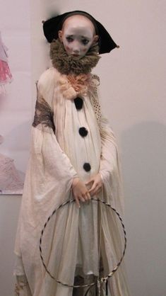 a creepy doll dressed in white and black is holding a metal hoop while standing next to a wall