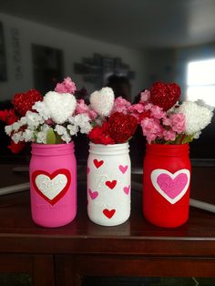 three painted mason jars with hearts and flowers in them on top of a wooden table