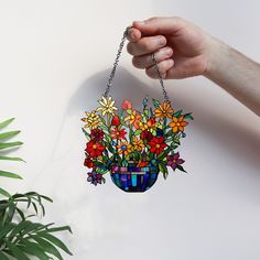 a hand holding a colorful flower pot hanging from a chain on a white wall next to a houseplant