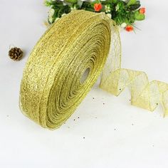 a roll of gold glitter ribbon next to a pine cone on a white surface with flowers in the background