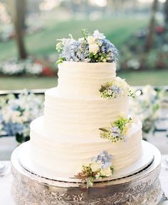 a white wedding cake with blue and white flowers on the top is sitting on a silver platter