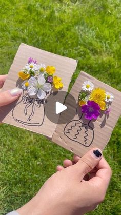 someone holding up two pieces of paper with flowers in them on top of the grass