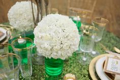 white flowers in green vases on a table with gold rimmed glasses and place settings