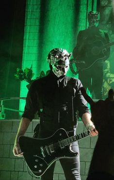 a man wearing a mask and holding a guitar in front of a green light with other people behind him