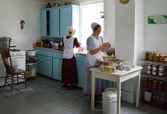 two women in aprons are preparing food in an old fashioned kitchen with blue cabinets