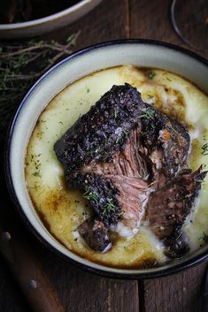 a bowl filled with meat and potatoes on top of a wooden table next to utensils