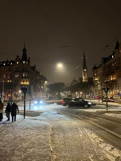 two people walking down a snowy street in the city at night with cars passing by