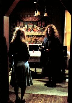 two women standing in a kitchen talking to each other and looking at something on the table