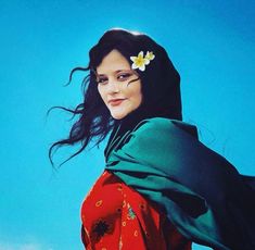 a woman with long hair wearing a green shawl and flower in her hair, against a blue sky