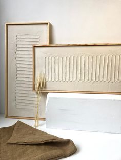 two framed art pieces sitting next to each other on a white table top with a brown cloth in front of them