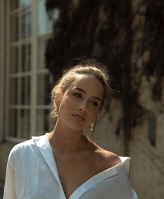 a woman with long hair wearing a white shirt and gold hoop earrings standing in front of a building