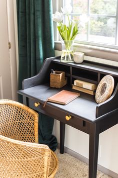 a desk with a book, vase and flowers on it in front of a window