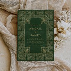 a green and gold wedding card sitting on top of a white cloth next to flowers