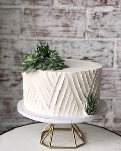 a white cake with succulents on top sitting on a stand next to a brick wall