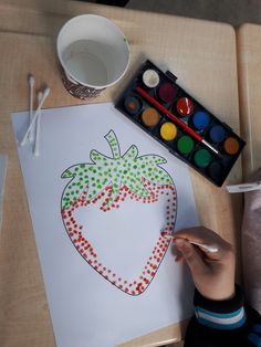 a child's hand is holding a paintbrush and drawing a strawberry on paper