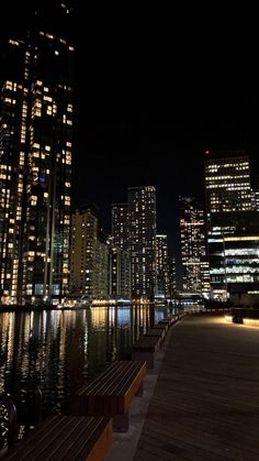 the city skyline is lit up at night with lights reflecting in the water and benches along the waterfront