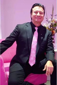 a man in a suit and tie sitting on a pink chair with flowers behind him