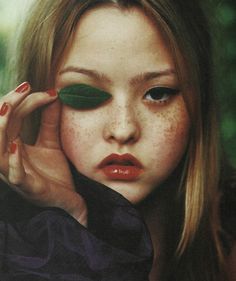 a girl with freckled hair and green leaves on her eyes, holding onto one leaf