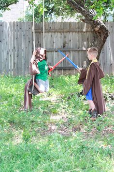 two children dressed up in costumes playing with lightsabens on the grass near a tree