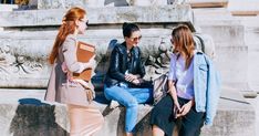 three women are sitting on a stone wall talking to each other and one is holding a book