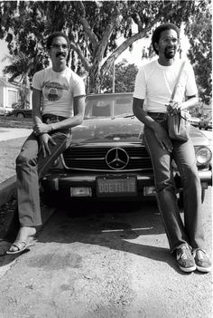 two men sitting next to each other on the side of a road in front of a car