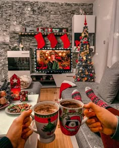 two people holding coffee mugs in front of a christmas tree with stockings on it